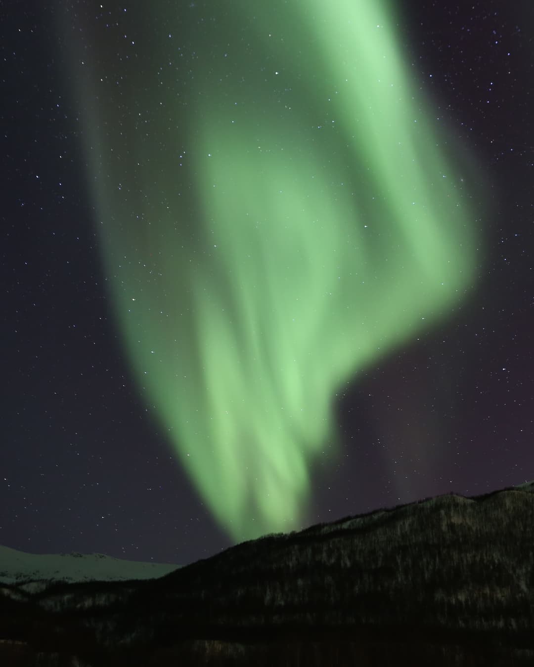 Una cortina de aurora verde brillante sobre una montaña oscura bajo un cielo estrellado cerca de Tromsø, norte de Noruega.
