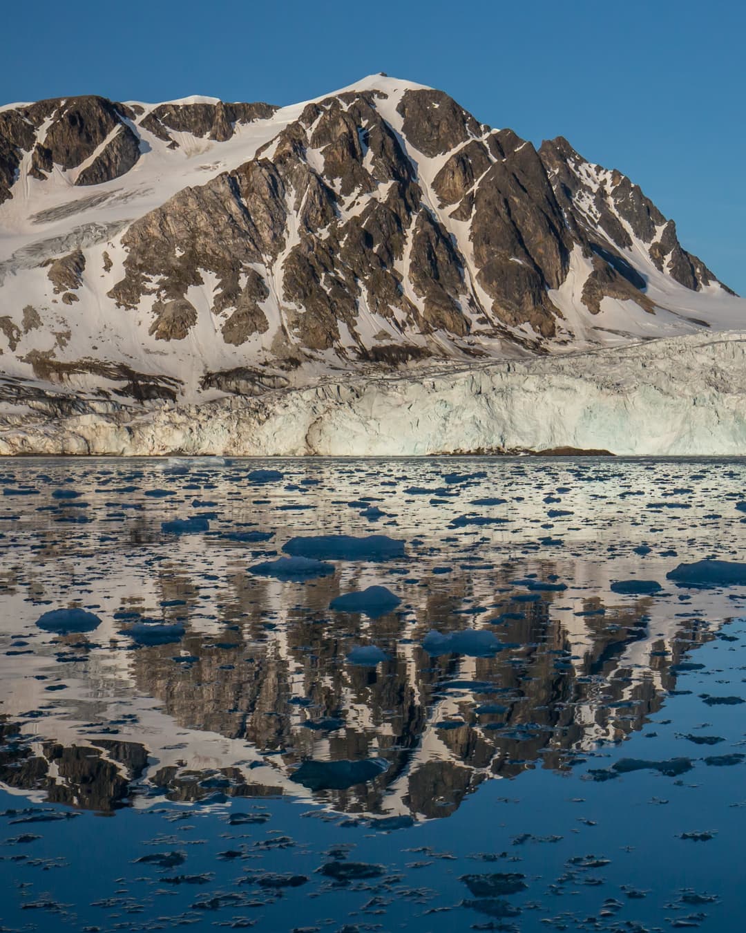 Un frente glaciar bajo picos nevados, reflejado en un fiordo en calma salpicado de hielo flotante, en el noroeste de Spitsbergen, Svalbard.